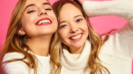 Portrait of two young stylish smiling blond women. Girls dressed in summer hipster clothes. Positive models making selfie on smartphone on pink background Beneficios de ortodoncia en adultos